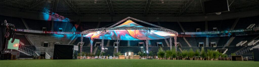 Large event tent set up in stadium for concert or festival, illuminated with colorful stage lighting.