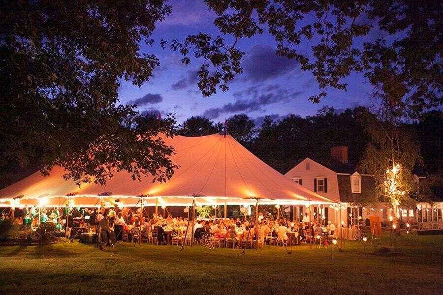 Elegant outdoor tent setup for wedding reception in backyard, illuminated with string lights at dusk.