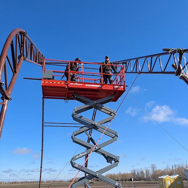 Elevated view of workers on a cherry picker working on a tall amusement park ride against a bright blue sky.